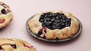 A rustic blueberry galette with juices dribbling down one side in a grey pie plate on a pink background, with two cropped lattice-top blueberry pies in the background