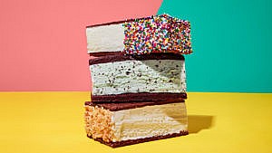 A stack of ice cream sandwiches, with different types of ice cream, on chocolate wafers; one is dipped into sprinkles, one into coconut