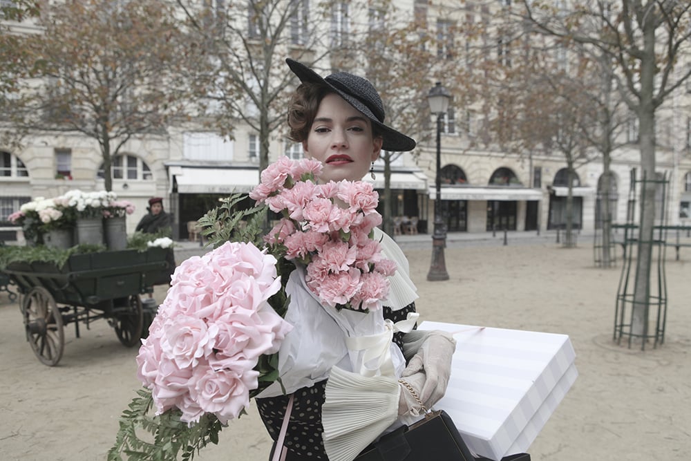 Lily James against a Paris street backdrop wearing a dramatic tilted hat and holding bunches of pink flowers