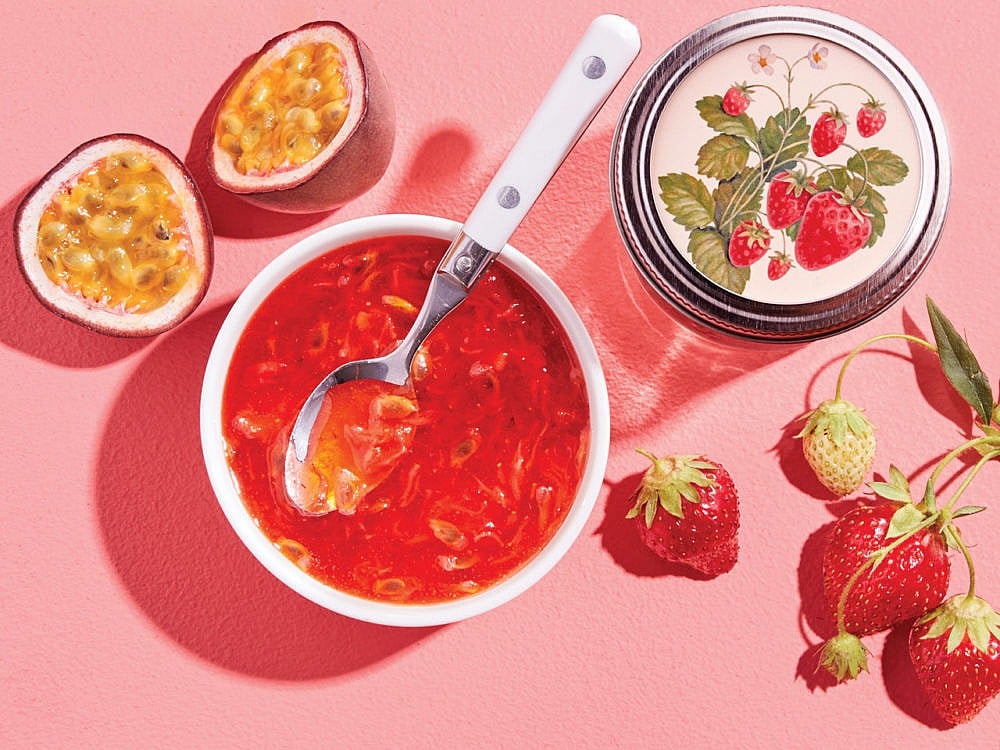 A split open passion fruit sits next to a bowl of jam with a spoon in it, a jam jar with strawberries on it, and a group of 4 strawberries against a pink background