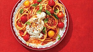 A plate of buccatini with cherry tomatoes and burrata against a red background