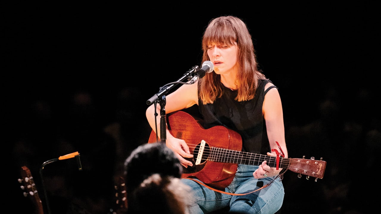 Leslie Feist, seated, holding a guitar and singing into a microphone