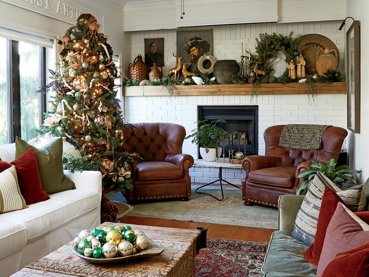 A Christmas tree in front of a whitewashed brick fireplace, with two brown leather armchairs and a coffee table with a bowl of Christmas ornaments in the foreground