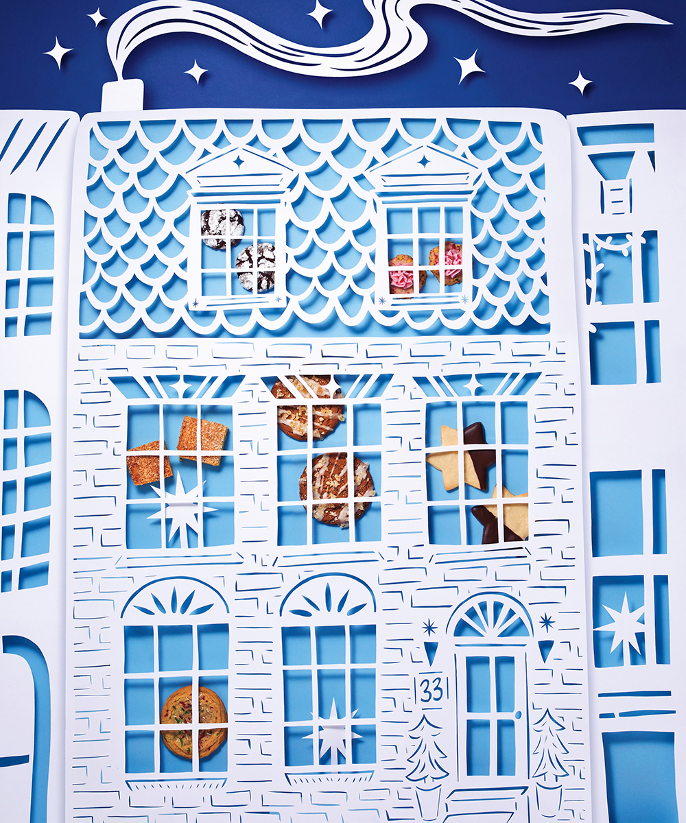A paper-art cut out version of a holiday home, cut out of white paper, with six cookies in the post all behind the windows, on a blue background