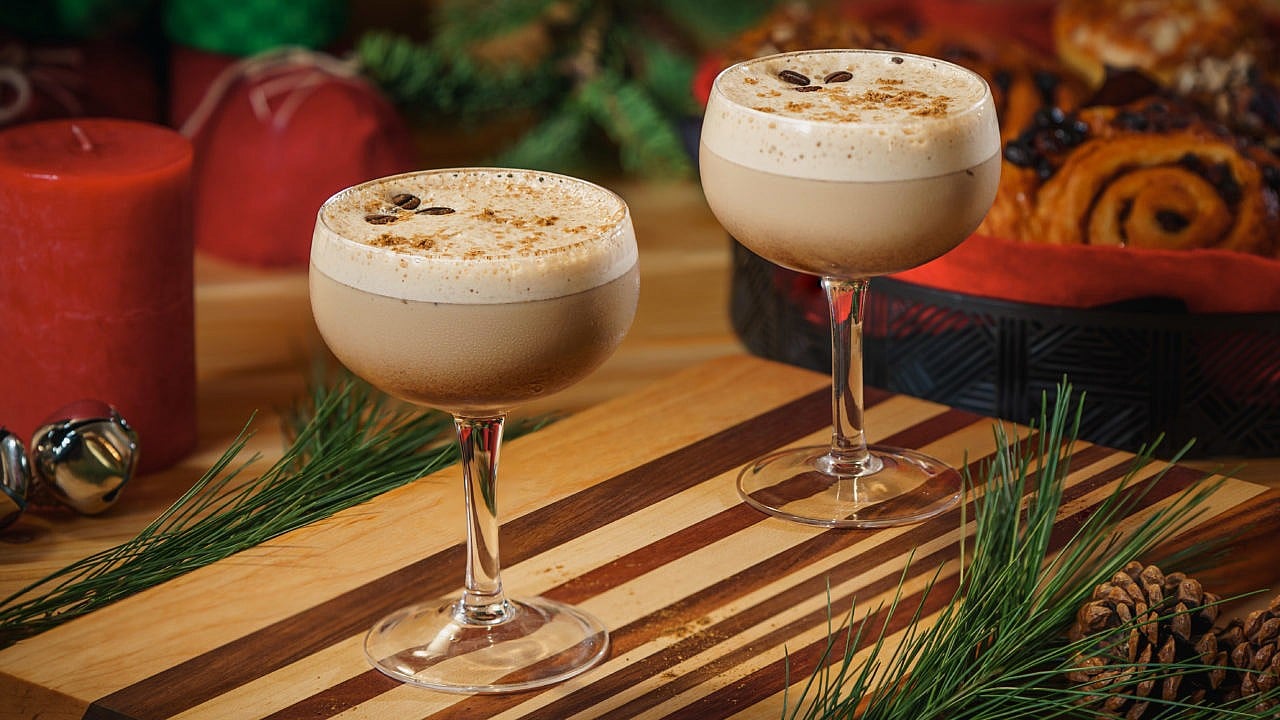 A pair of coupe glasses with a creamy, coffee-bean decorated cocktail inside, on a striped wood plant, surrounded by red candles and seasonal garnishes
