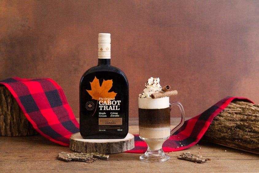 A bottle of Cabot Trail Maple Cream next to a three-layer coffee cocktail topped with whipped cream and a cinnamon stick on a buffalo check table runner