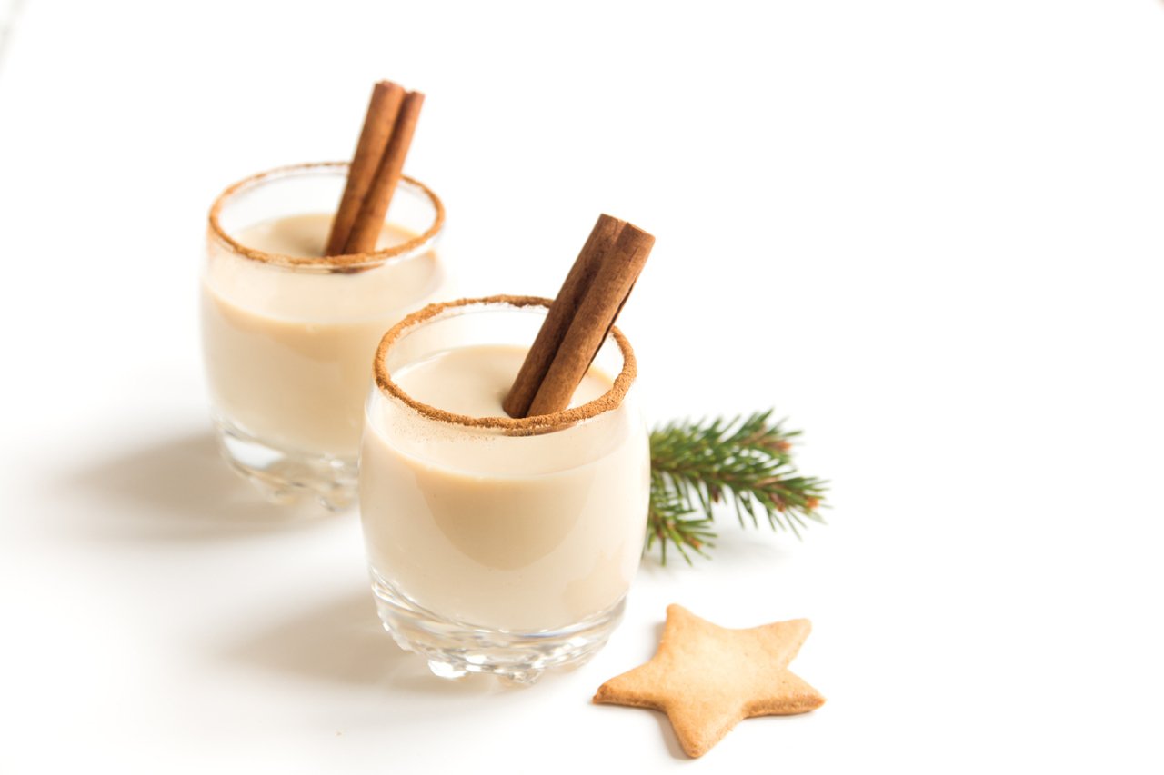Two small glasses of a cream coloured eggnog with a cinnamon stick in each glass