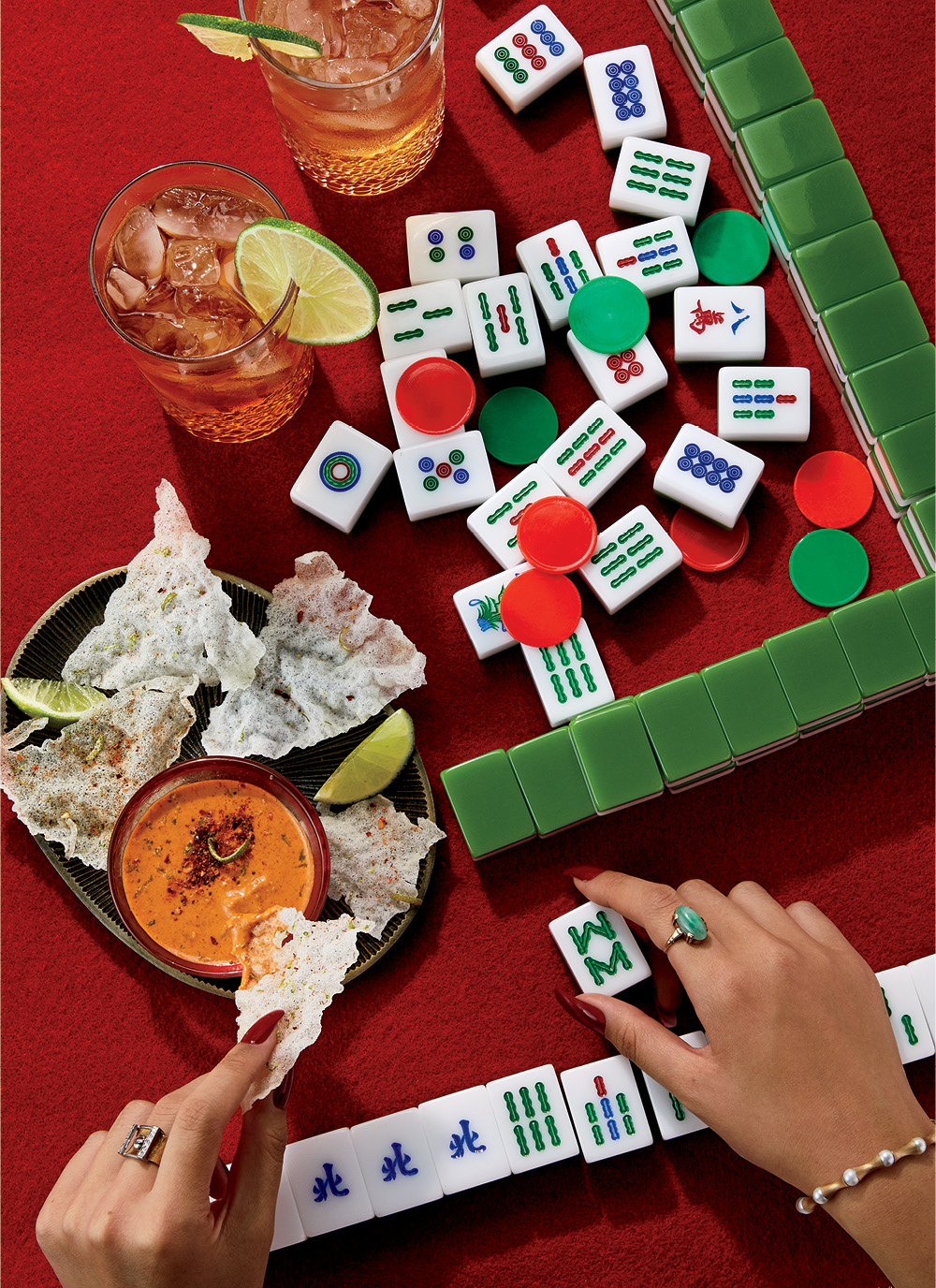 A table with mahjong tiles on a red tablecloth, you can see a woman's hands, she is eating from a bowl of rice paper chips with peanut dipping sauce, there are two spiced rum and ginger beer cocktails