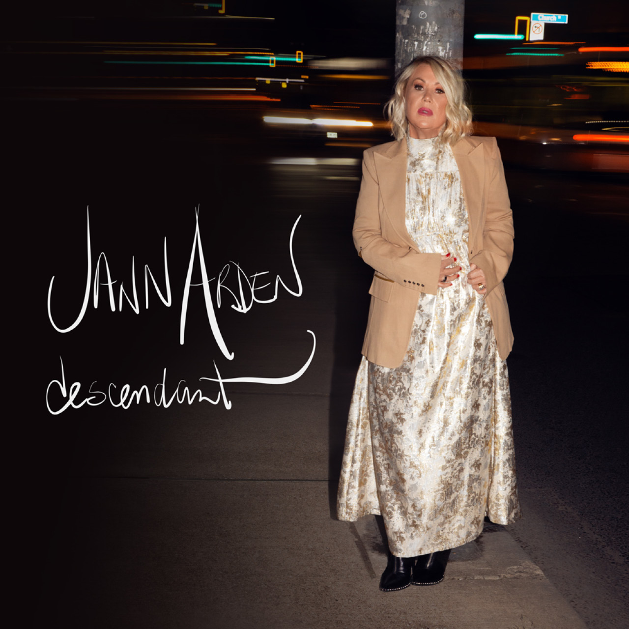 Standing on the sidewalk at nighttime, Jann Arden wears a long white dress with subtle detailing and a cream blazer.