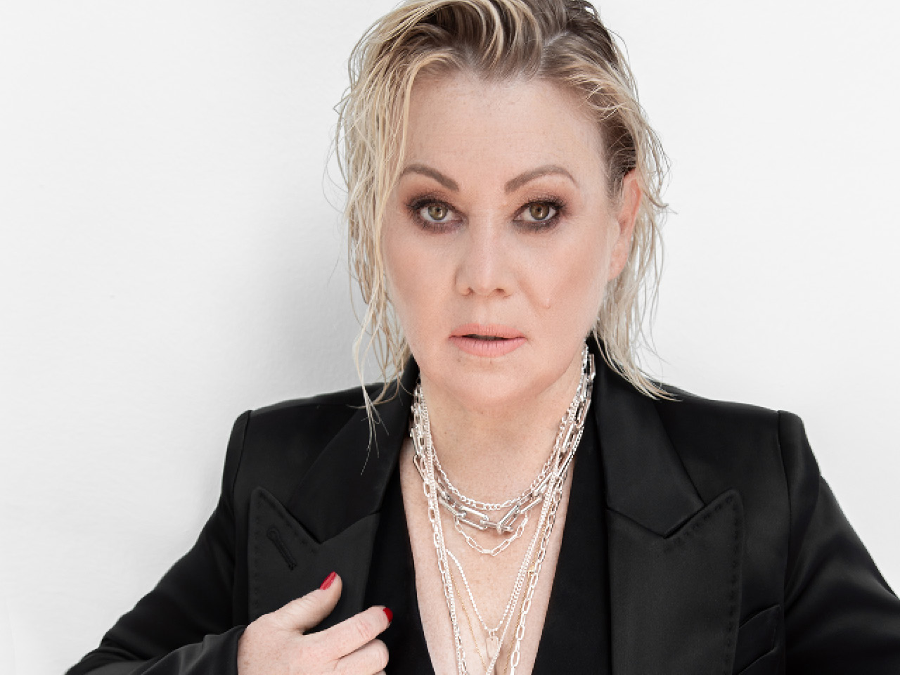 Jann Arden looking into the camera, standing in front of a white background wearing a black blazer and layered chain necklaces.