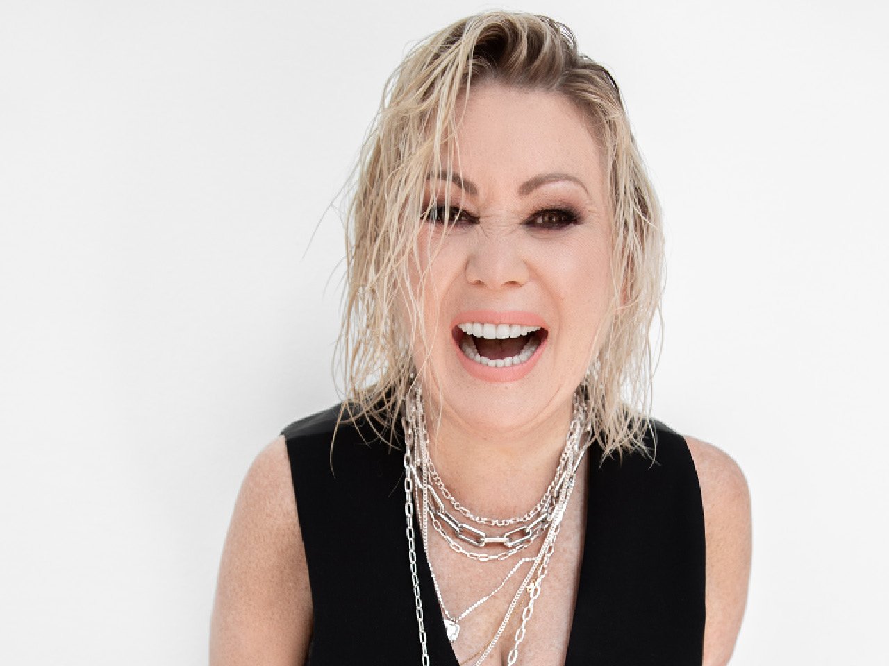 Jann Arden laughing in front of a white background wearing a black blazer and layered chain necklaces.