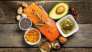 A filet of salmon, half an avocado, a small bowl of olives, a bowl of omega oil capsules and a few nuts sit on a wooden countertop.