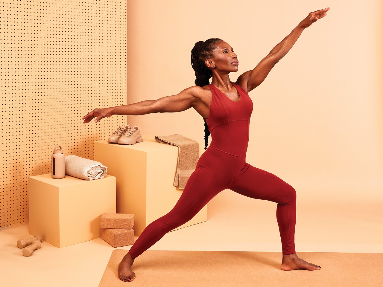 A woman doing yoga while wearing a red one-piece bodysuit against a sand-coloured background.
