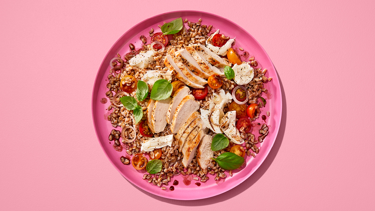 A pink bowl of farrosalad with chicken and caprese on a pink table.