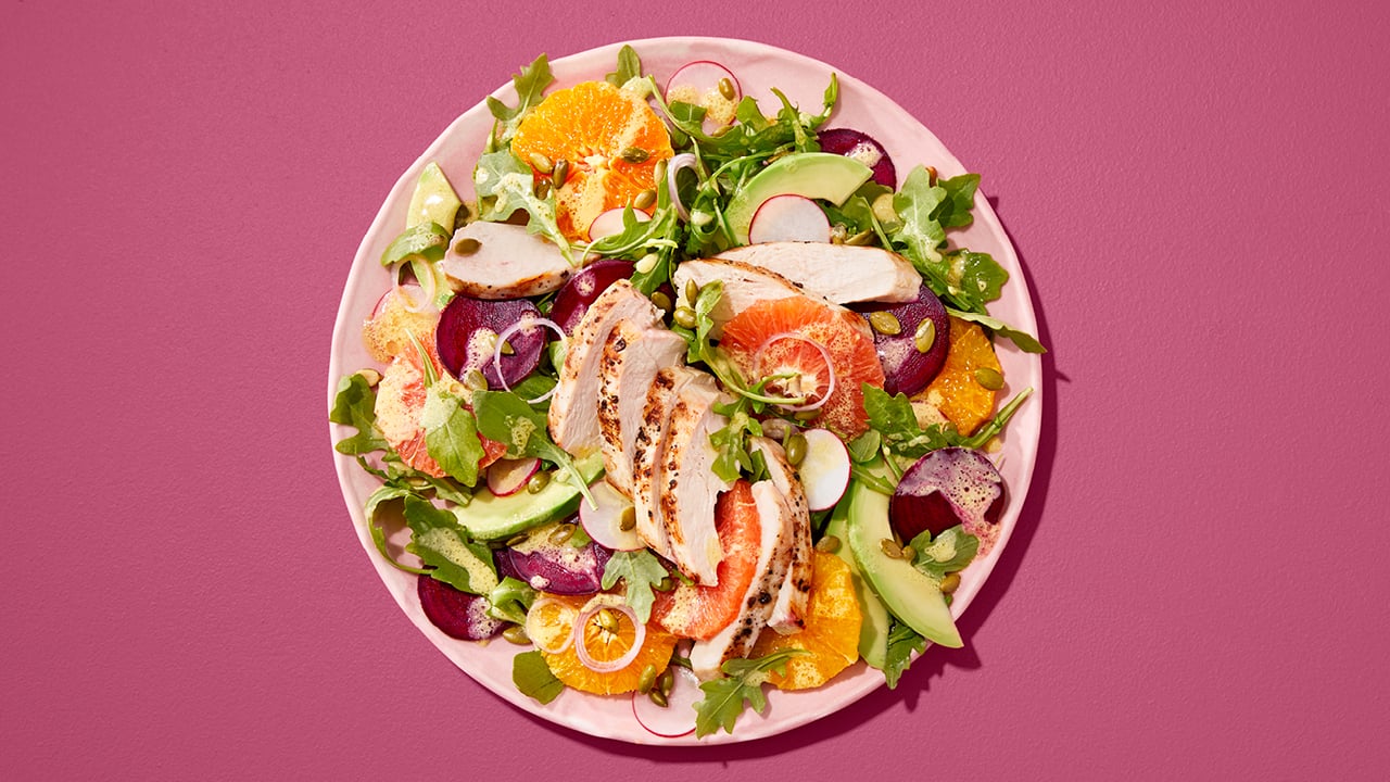 Salad with avocado, sliced ​​chicken and oranges on a pink plate in front of a purple background.