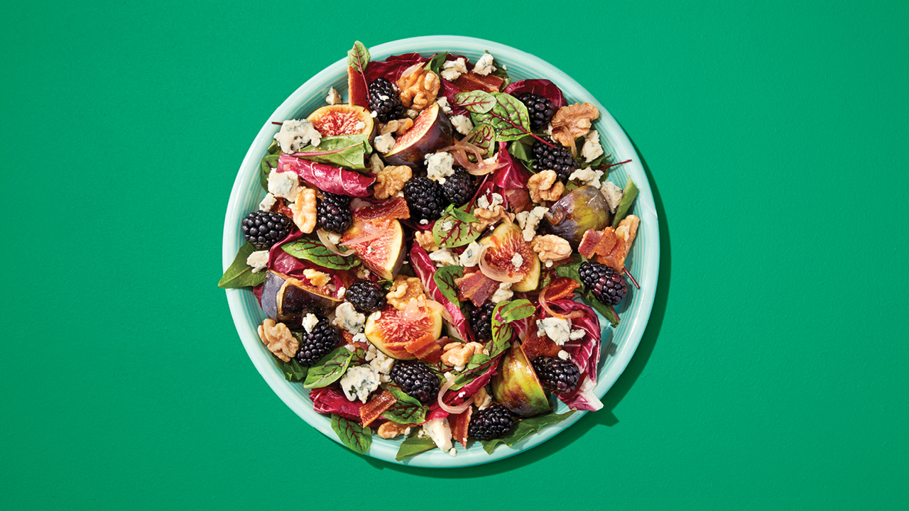 A salad with blackberries, figs, and greens on a green plate on a green table.