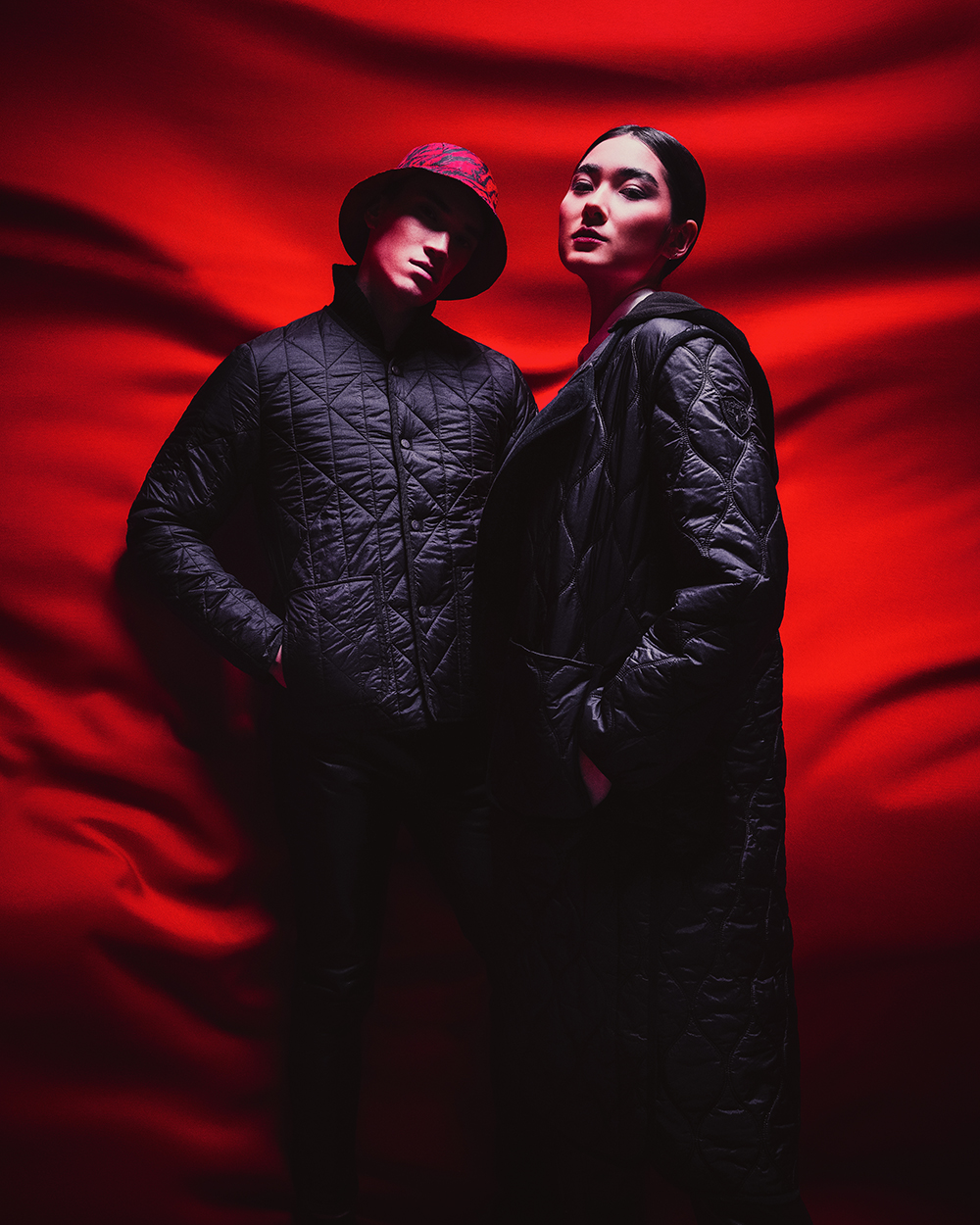 Two people in black jackets stand and look forward, against a red and black background.
