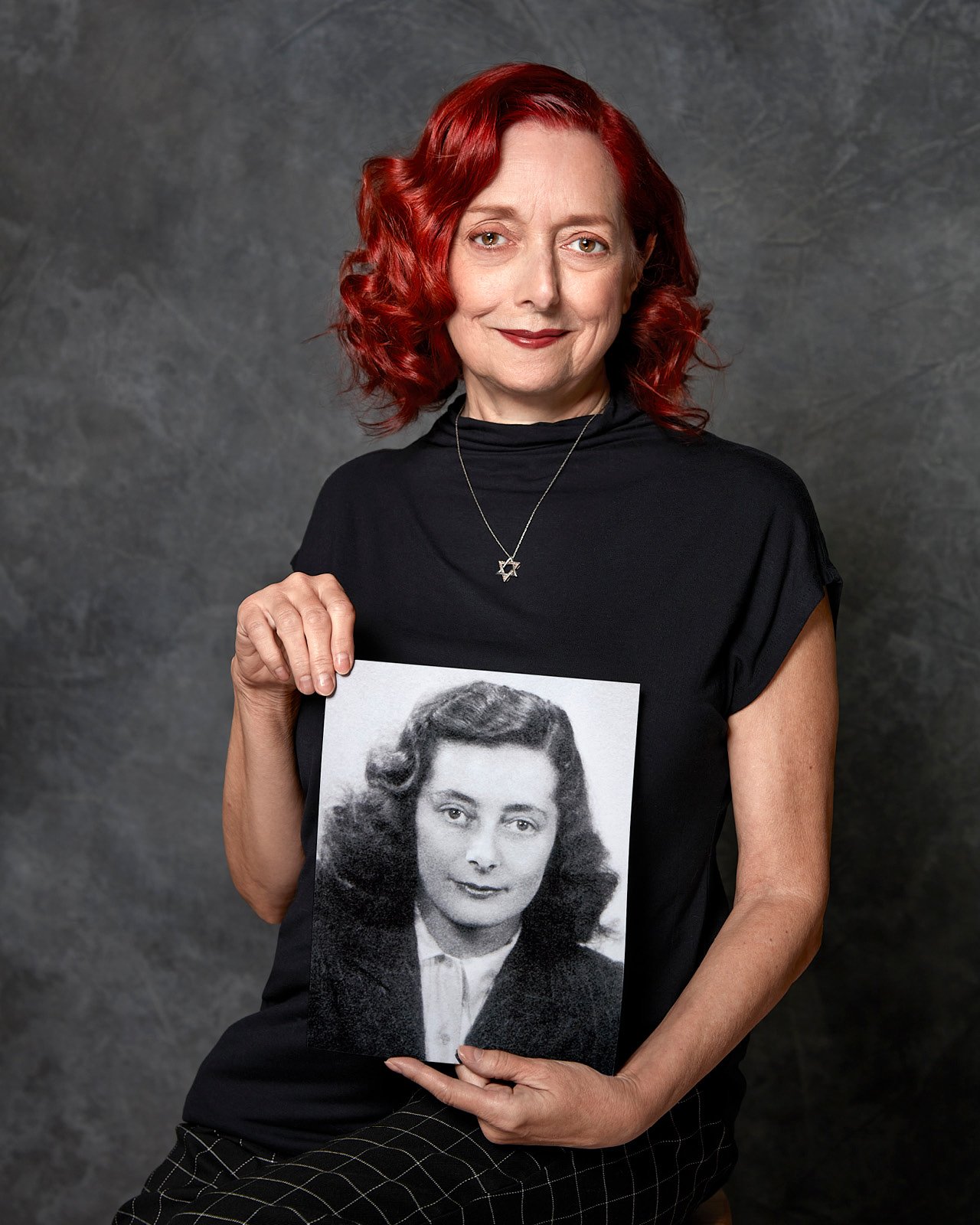 A woman with red hair sits against a grey background holding a black and white photograph of her mother.