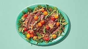 A plate of arugula topped with steak and tomatoes, on a green plate on a green table.