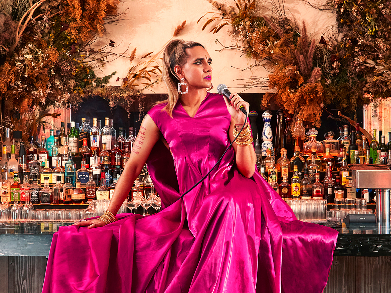 Author Vivek Shraya wearing a pink gown and holding a microphone while sitting on a bar.