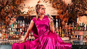 Author Vivek Shraya wearing a pink gown and holding a microphone while sitting on a bar.