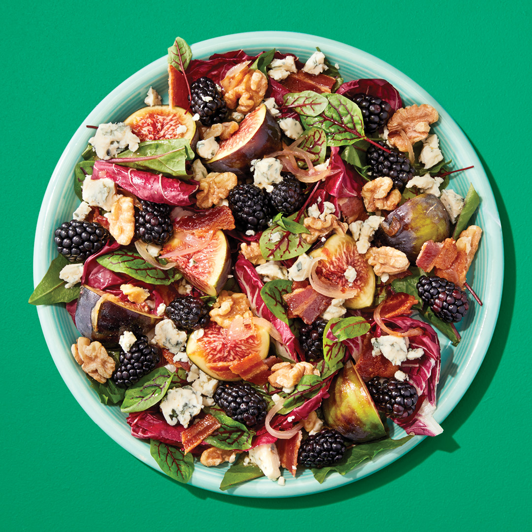 A salad with blackberries, figs, and greens on a green plate on a green table.