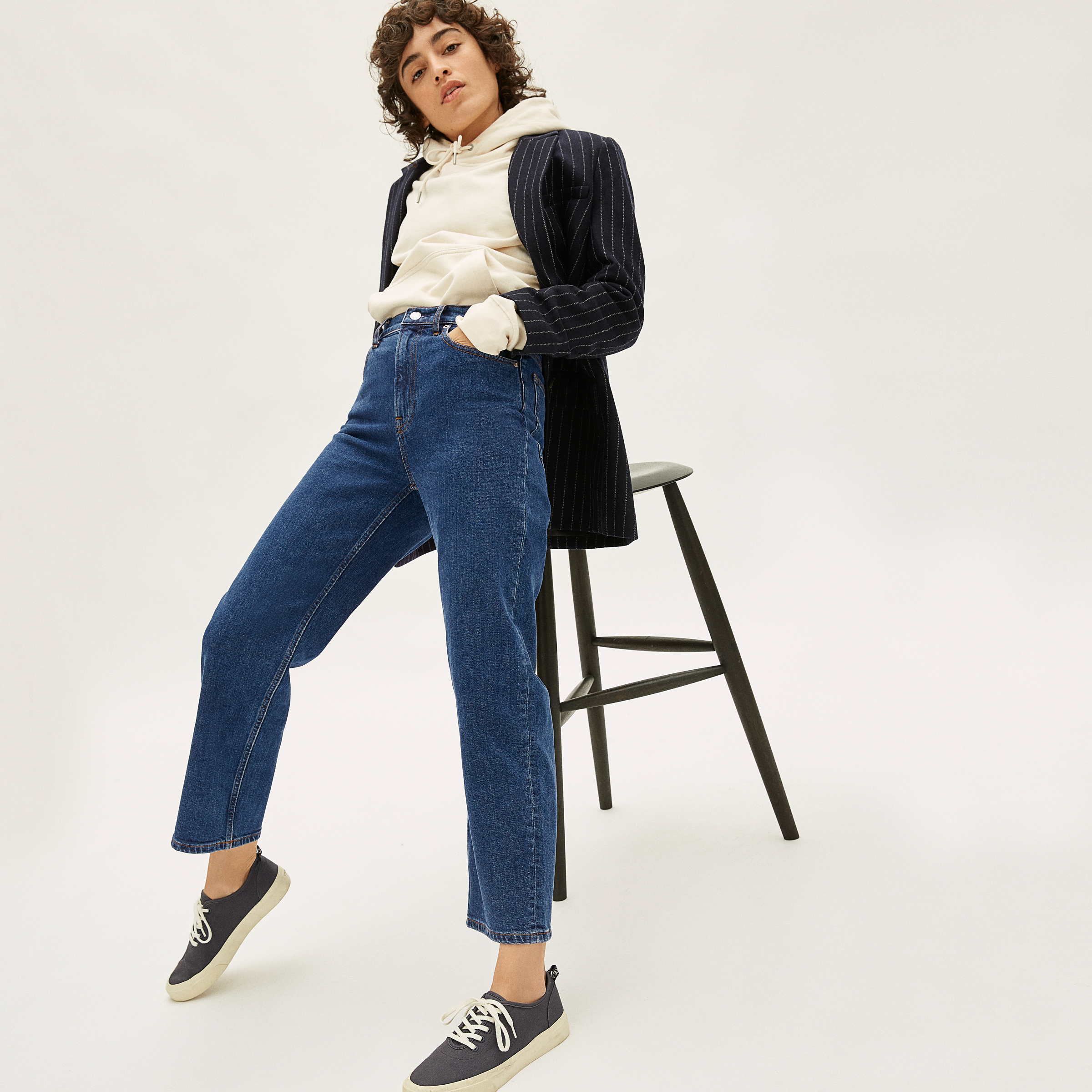 A model wearing dark blue jeans sits on a tall stool