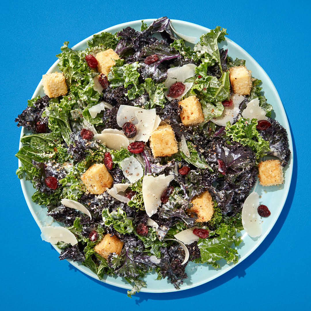 A ceasar salad with kale, anchovies and cranberries on a blue plate on a blue table.