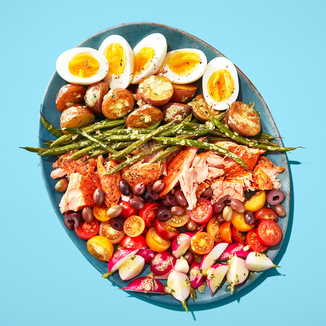 A blue plate with hard boiled eggs, green beans, olives, tomatoes on a blue table.