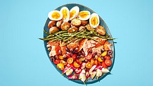 A blue plate with hard boiled eggs, green beans, olives, tomatoes on a blue table.