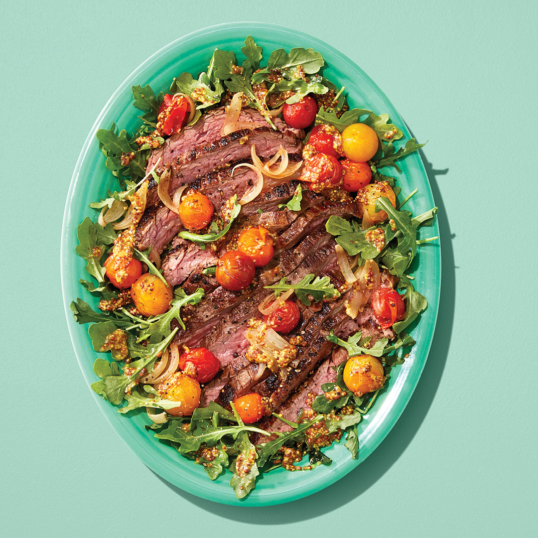A plate of arugula topped with steak and tomatoes, on a green plate on a green table.