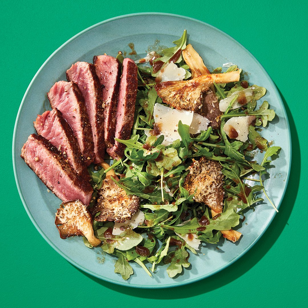 A salad with sliced beef, greens and mushrooms on a green plate on a green table.
