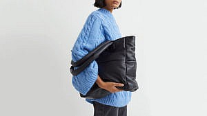 A model wearing a blue sweater and holding a black oversized padded handbag.