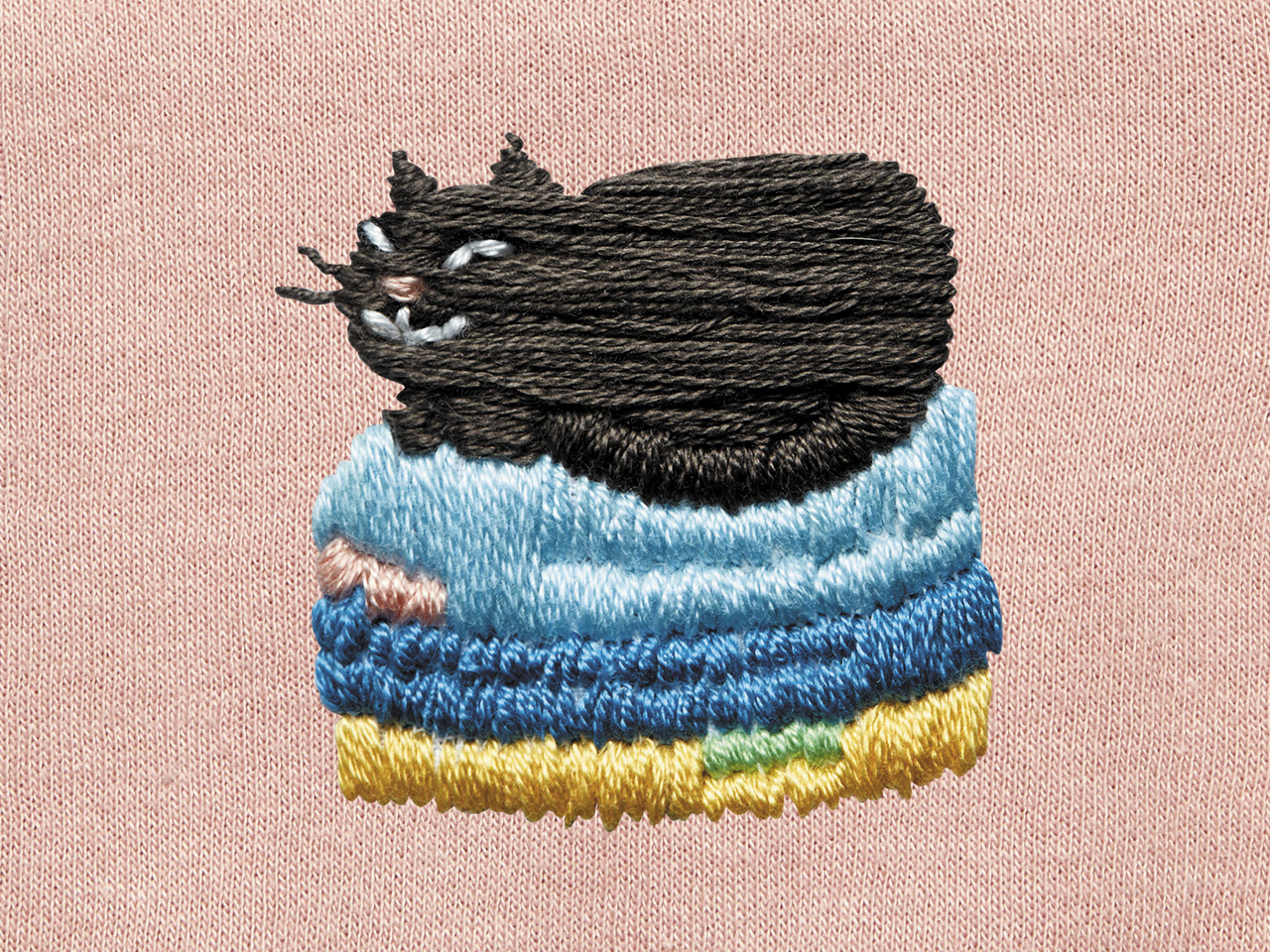 A photo of an embroidery of a black cat sitting on top of a laundry pile against a pink fabric background.