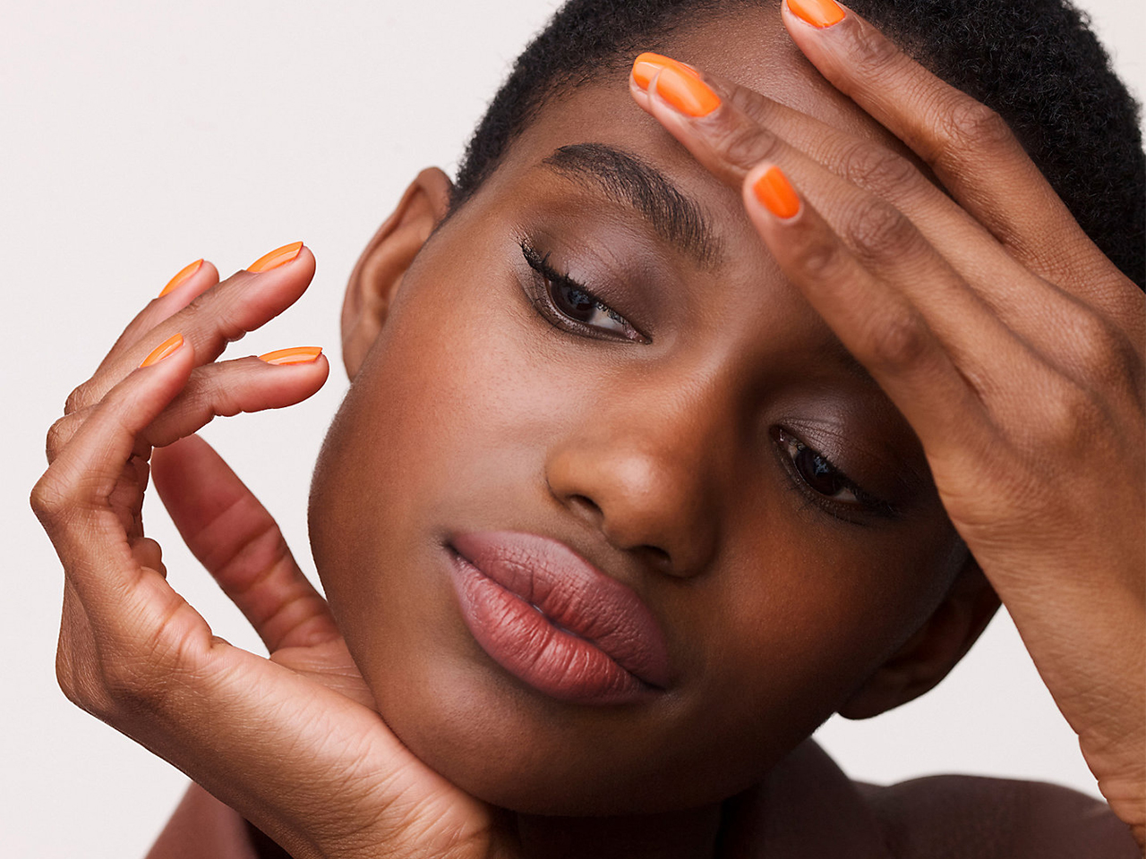 A model wearing orange nail polish to illustrate a round-up of the best nail polish colours for spring 2022.