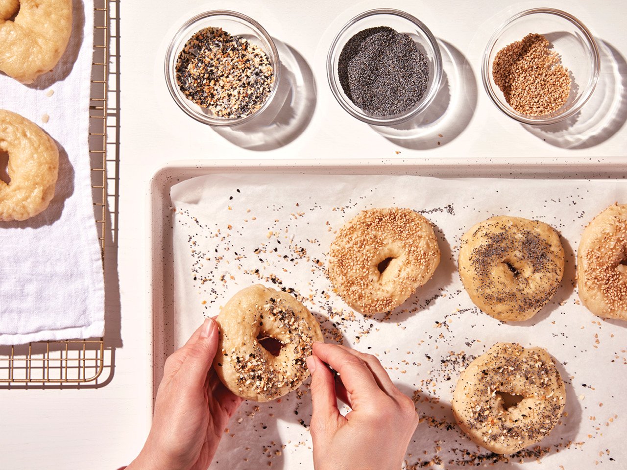 A photo of a person's hands covering homemade bagel in a mixture of sesame and poppy seeds