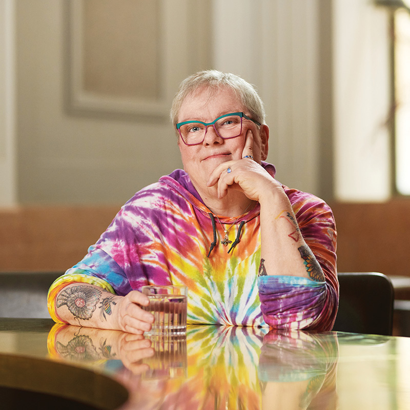 Older woman sitting at a counter, holding a mug wearing a tye die sweater, and glasses. 