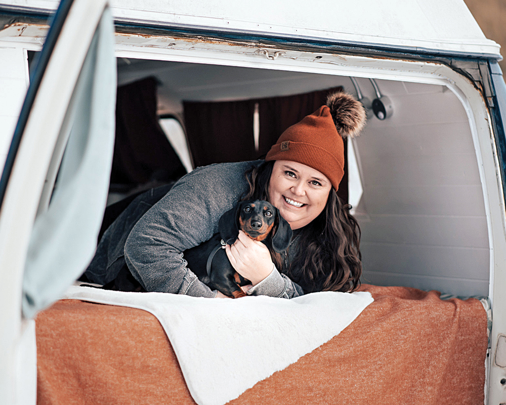 Photo of Emily Inson holding her dog in the back of her van with a brown tuque on and grey longsleeve.