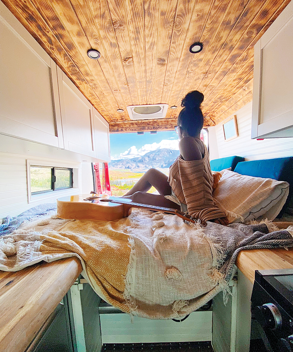 Joanie sitting on her bed in her van, with a brown guitar next to her and a scenic view. 
