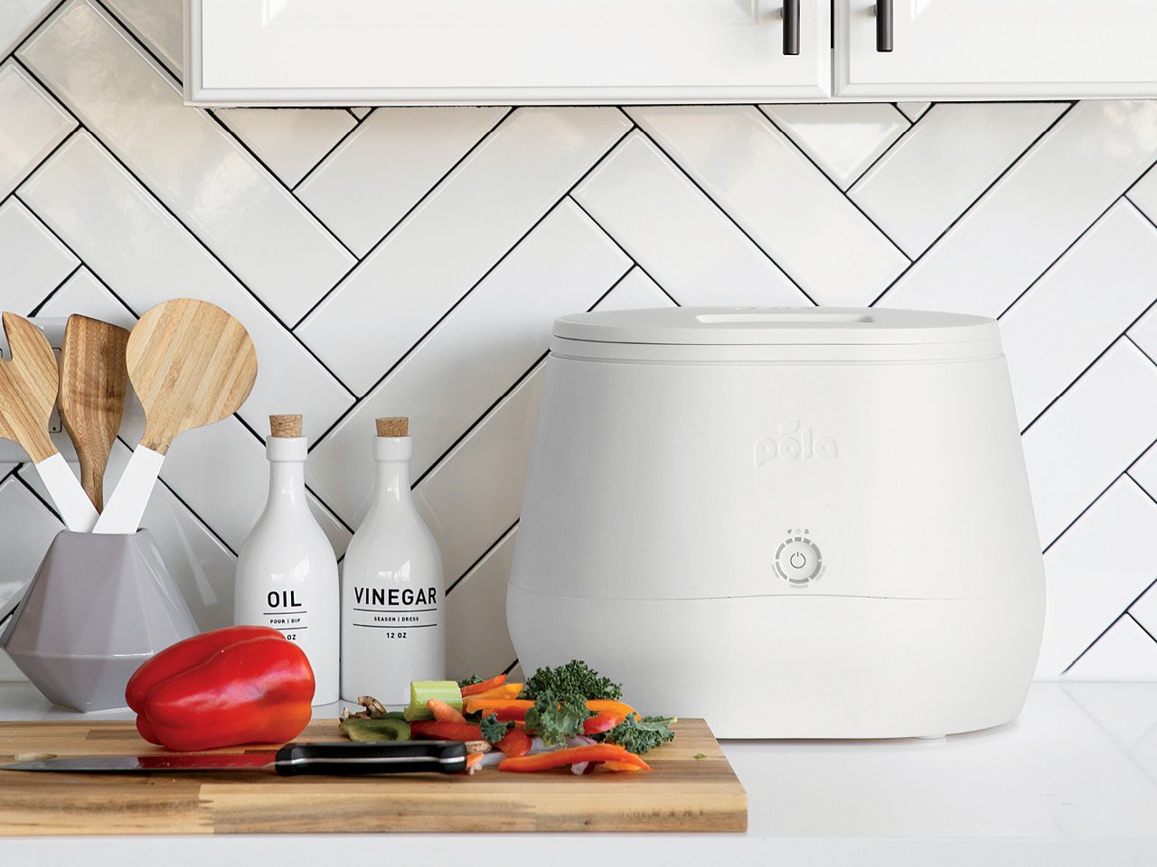 A white countertop composter from LOMI on a kitchen counter beside condiments, cooking tools, and a chopping board