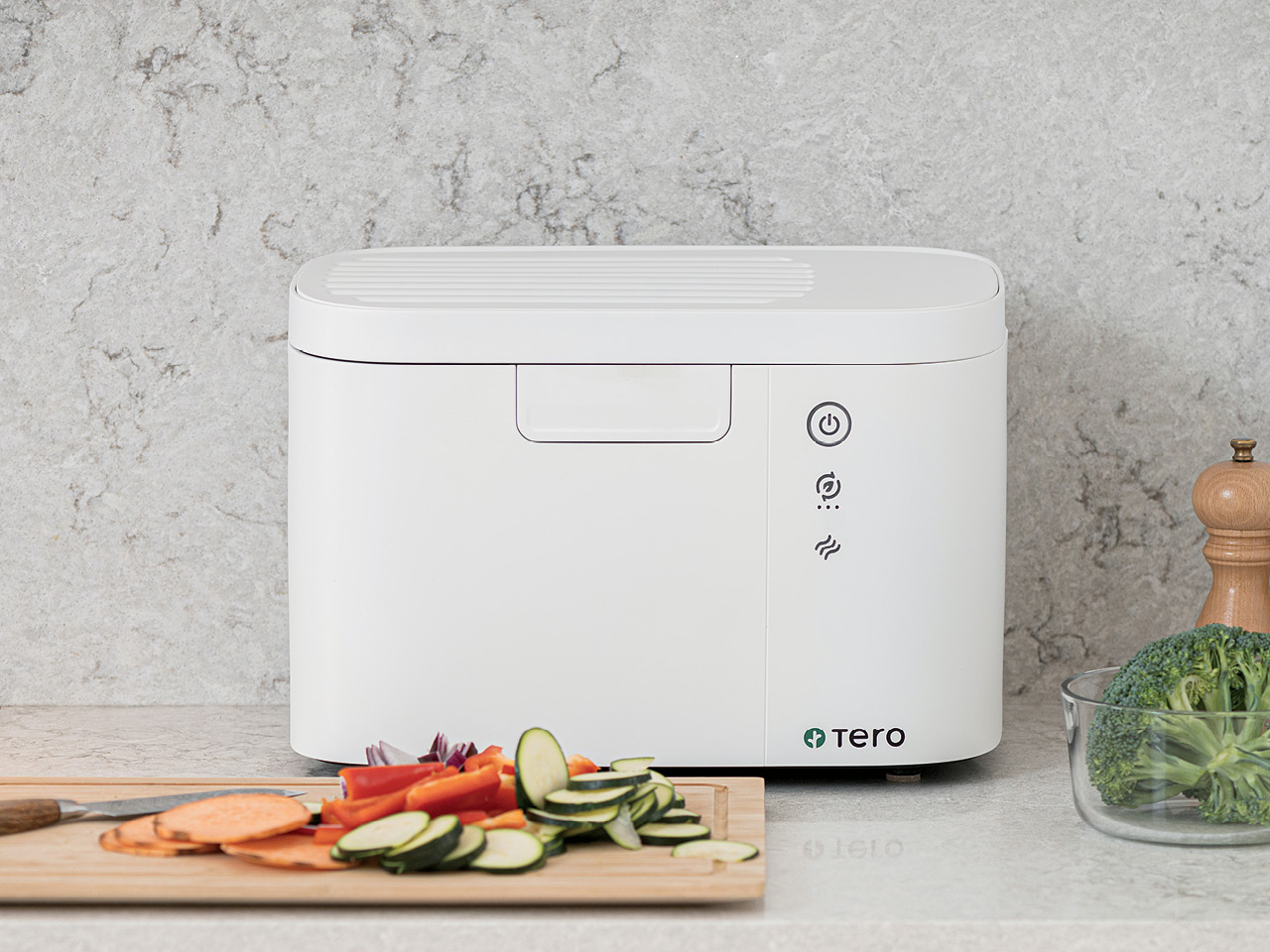 A white countertop composter from TERO positioned on a kitchen counter with sliced vegetables