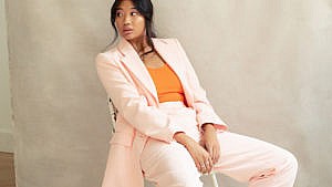 A model sitting on a chair wearing a pale pink blazer and pants and orange top for a round-up of dopamine dressing pieces for summer.
