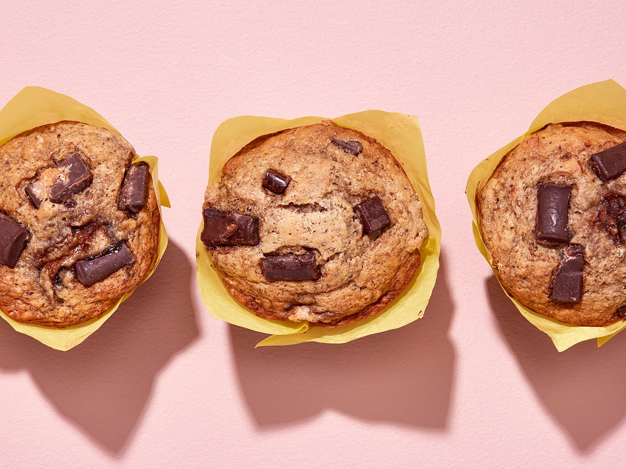 Three vegan banana chocolate chunk muffins in yellow wrapper on a pink background