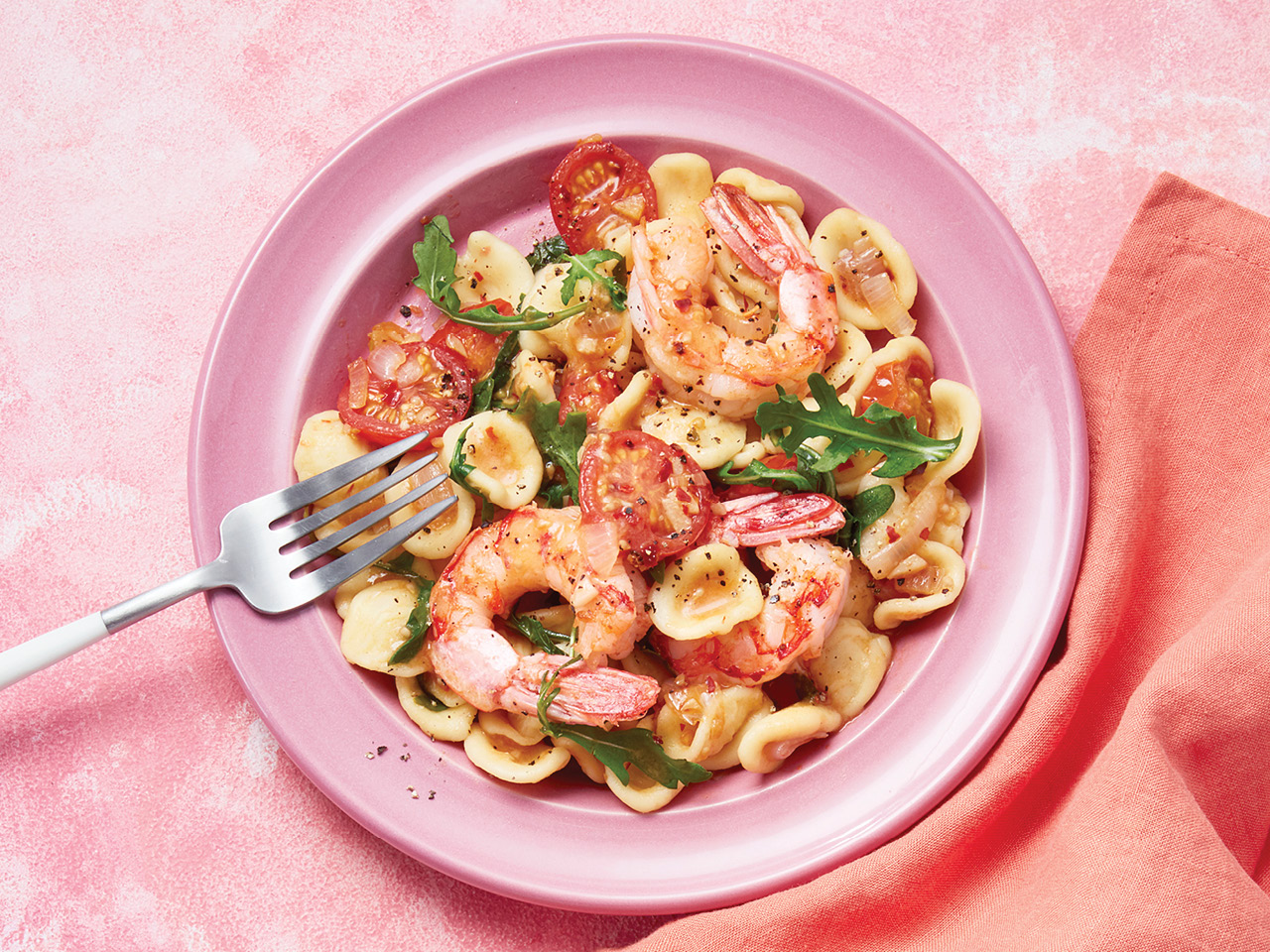 Pasta, shrimp, tomato and arugula in a white bowl on a pink background with a used spoon beside.