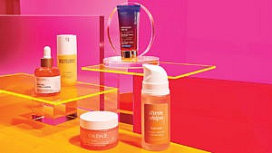 An array of packaged beauty products, including a tube of makeup, a vial of cream, and more, on clear stands against a sunset-hued, pink and orange background