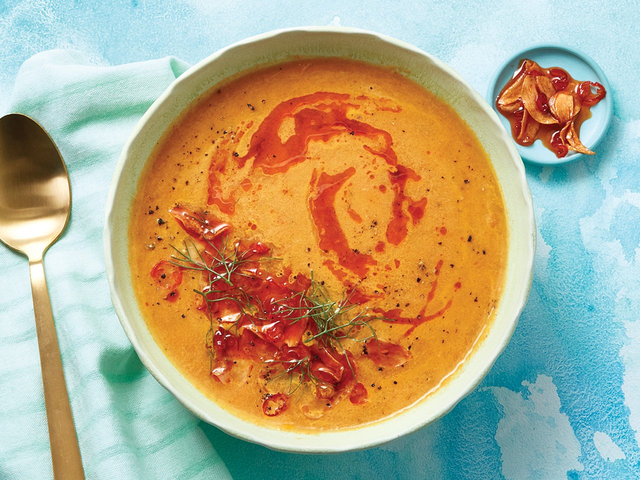 Carrot fennel soup with garlic chili oil on top in a light green bowl and a cloud printed background and along with a spoon.