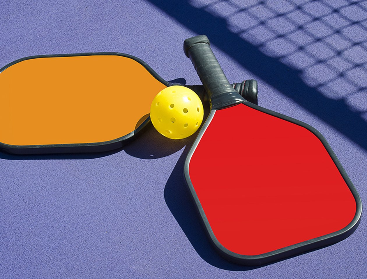 Image of 2 Pickleball paddles and a pickleball laying on pickleball court with net shadow.