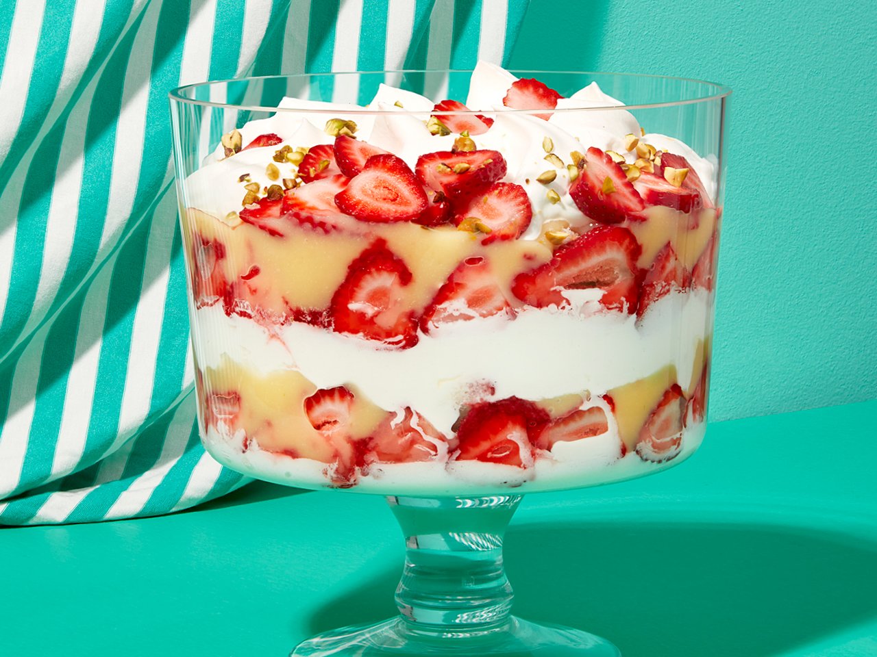 A trifle bowl showing layers of strawberry and lime curd trifle with pistachios on a turquoise table with a striped qhite and turquoise curtain in the background