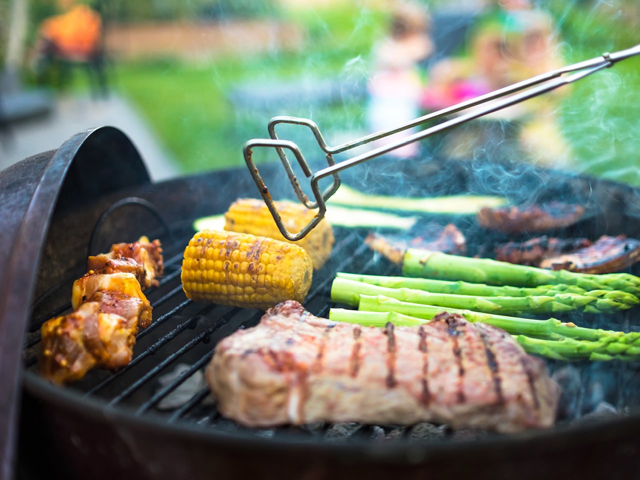 A pair of silver tongs suspended over an open black grill with various meats and vegetables grilling.