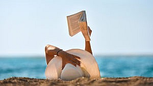 A woman reads a book on the beach wearing a giant sun hat